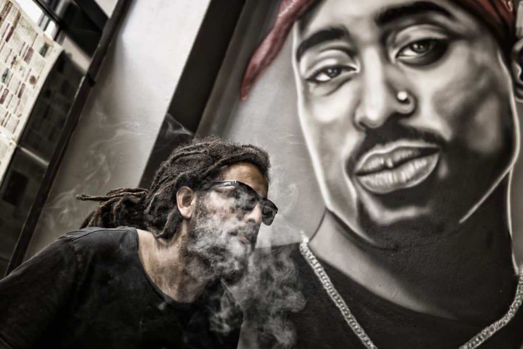 Man with dreadlocks and sunglasses poses near Tupac portrait